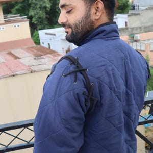 Rear three-quarter view of a man wearing a blue quilted medieval coat showing black cord lacing details at the shoulder.