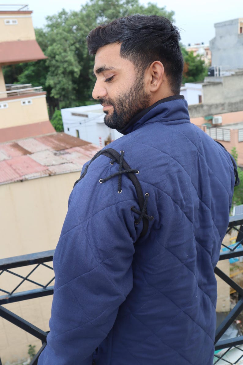 Rear three-quarter view of a man wearing a blue quilted medieval coat showing black cord lacing details at the shoulder.