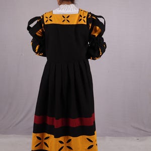 Pode incluir: Um vestido preto com detalhes em amarelo e bord&ocirc;. O vestido apresenta um decote quadrado com uma gola branca com babados e recortes decorativos. As mangas s&atilde;o franzidas nos pulsos. A saia tem listras horizontais amarelas e bord&ocirc;.