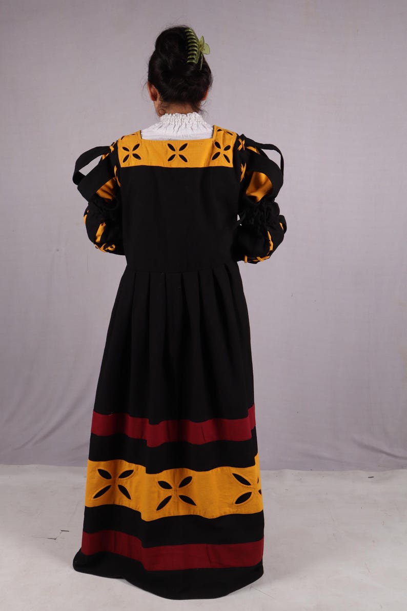 Pode incluir: Um vestido preto com detalhes em amarelo e bord&ocirc;. O vestido apresenta um decote quadrado com uma gola branca com babados e recortes decorativos. As mangas s&atilde;o franzidas nos pulsos. A saia tem listras horizontais amarelas e bord&ocirc;.