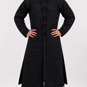A smiling male model wearing a black quilted long gambeson coat and brown boots, standing with hands on hips against a white background.