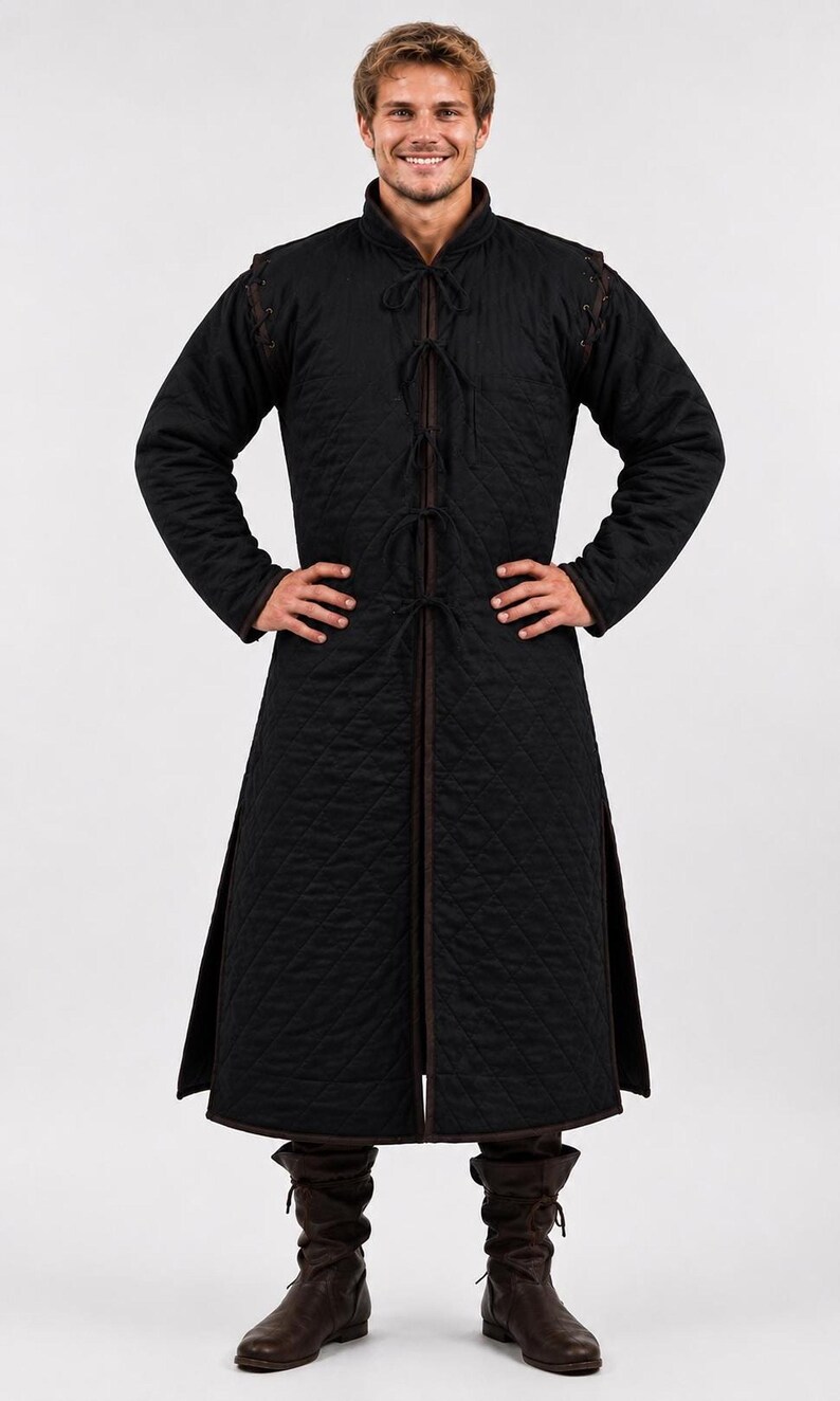 A smiling male model wearing a black quilted long gambeson coat and brown boots, standing with hands on hips against a white background.