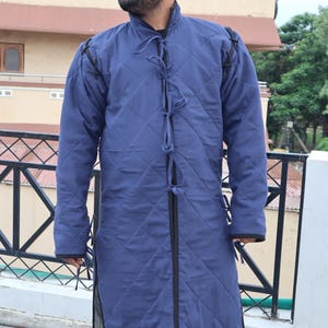A man wearing a long, blue quilted medieval gambeson coat with black binding and front tie closures while standing on an outdoor balcony.