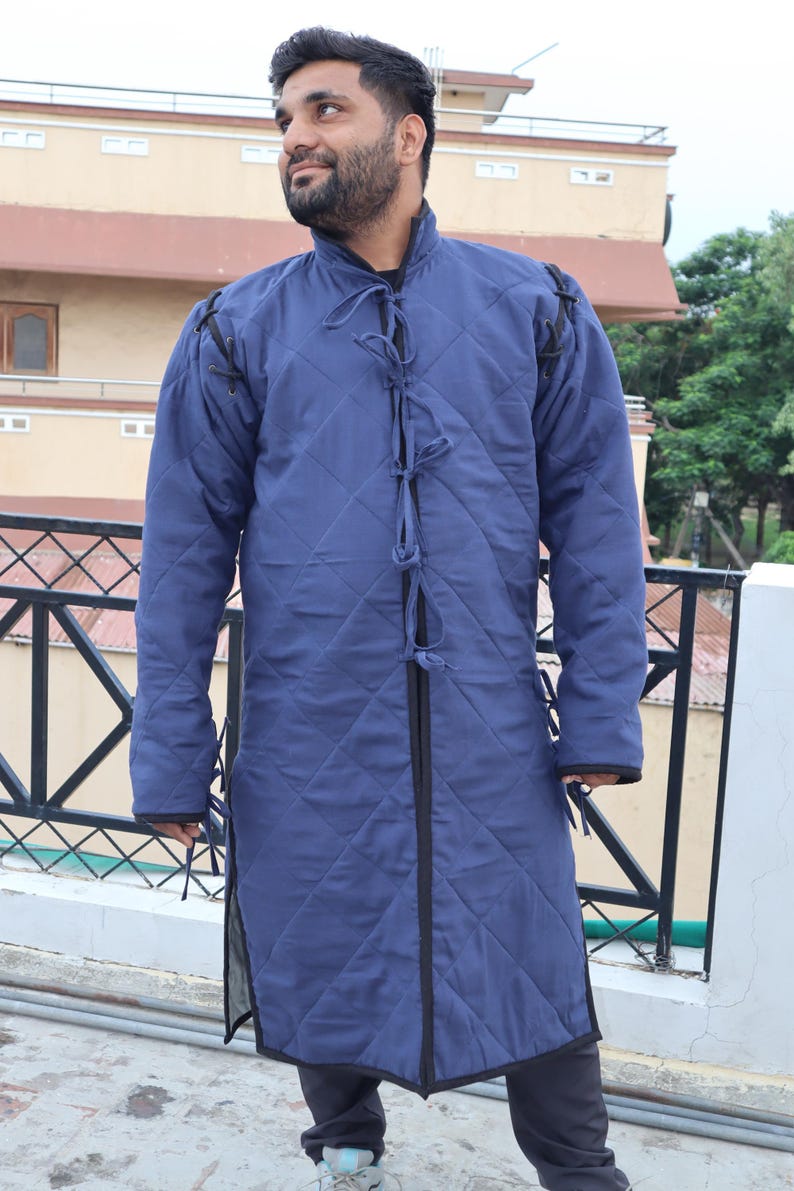 A man wearing a long, blue quilted medieval gambeson coat with black binding and front tie closures while standing on an outdoor balcony.