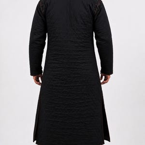 The back view of a man wearing a black quilted long gambeson coat and brown boots.