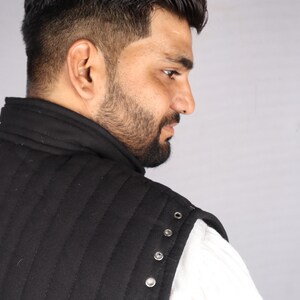 A close-up side view of a bearded man showing the shoulder seam of a black quilted gambeson with silver metal grommets.