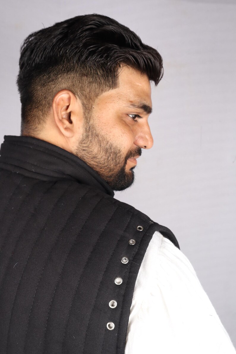 A close-up side view of a bearded man showing the shoulder seam of a black quilted gambeson with silver metal grommets.