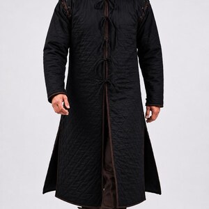 A man wearing a black quilted long gambeson coat walking forward toward the camera.