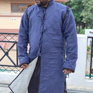A man wearing a blue quilted long gambeson coat, holding open the front hem to reveal a light-colored inner lining.