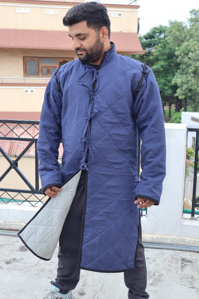 A man wearing a blue quilted long gambeson coat, holding open the front hem to reveal a light-colored inner lining.