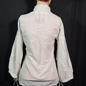 Pode incluir: Blusa branca de manga comprida com gola alta com babados e punhos franzidos. A blusa tem um corte solto e detalhes de cord&atilde;o na bainha e nos punhos. A pe&ccedil;a &eacute; exibida em um manequim.