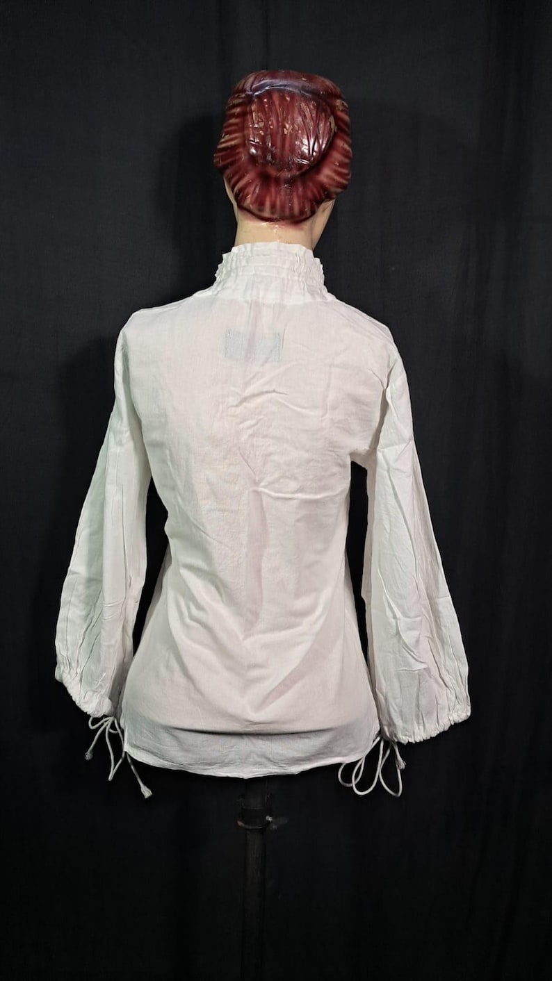 Pode incluir: Blusa branca de manga comprida com gola alta com babados e punhos franzidos. A blusa tem um corte solto e detalhes de cord&atilde;o na bainha e nos punhos. A pe&ccedil;a &eacute; exibida em um manequim.