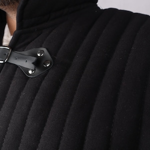 A close-up view of the collar and shoulder of a black quilted gambeson showing a leather strap closure and silver metal grommets.