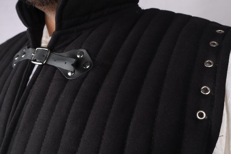 A close-up view of the collar and shoulder of a black quilted gambeson showing a leather strap closure and silver metal grommets.