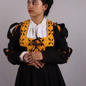 Pode incluir: Um vestido preto tradicional com um corpete amarelo e preto com um desenho floral. O vestido tem mangas compridas e uma blusa branca por baixo. As mangas t&ecirc;m punhos brancos. A mulher est&aacute; vestida com uma roupa tradicional.