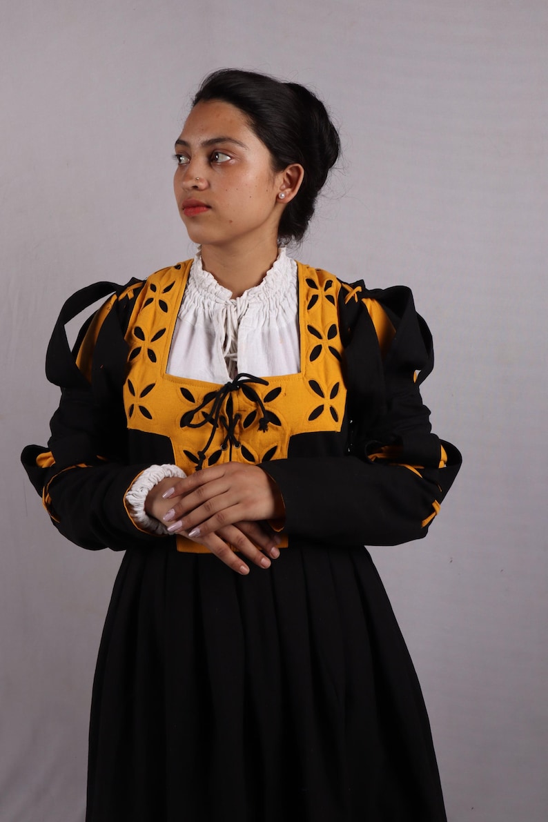 Pode incluir: Um vestido preto tradicional com um corpete amarelo e preto com um desenho floral. O vestido tem mangas compridas e uma blusa branca por baixo. As mangas t&ecirc;m punhos brancos. A mulher est&aacute; vestida com uma roupa tradicional.