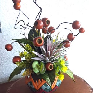 May include: A decorative arrangement featuring a round, patterned bowl with orange, teal, and dark red geometric designs. The bowl contains a mix of artificial greenery, light green succulents, and thin, dark brown branches tipped with round, reddish-brown berries.