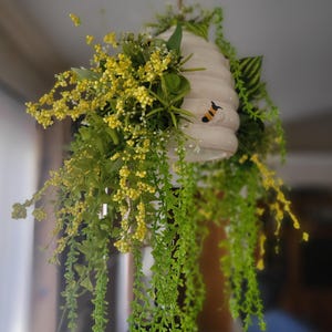 May include: A hanging decorative beehive with a painted bee and cascading greenery. The beehive is white with a textured surface. The greenery includes yellow and white flowers and trailing green vines.