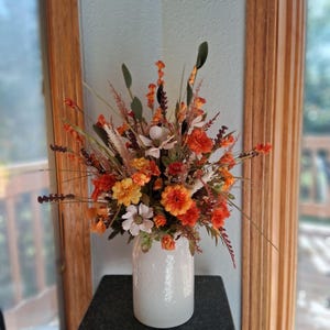 May include: A floral arrangement in a white cylindrical vase featuring a mix of artificial flowers and foliage in shades of orange, yellow, cream, and deep red. The bouquet includes various blossoms, tall grasses, and dark green leaves.