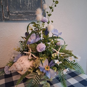 May include: A floral arrangement featuring light blue and lavender flowers, green foliage, and two white starfish. A large, textured beige seashell is nestled at the base among the stems, creating a coastal-themed decorative centerpiece.