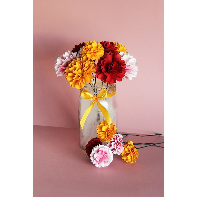 May include: A bouquet of paper flowers in shades of red, yellow, and white arranged in a clear glass jar with a yellow ribbon. The flowers are made of paper and have a realistic appearance.