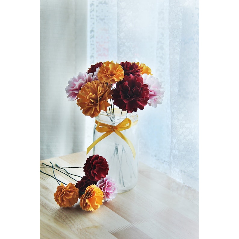 May include: A bouquet of paper flowers in shades of red, orange, and pink arranged in a clear glass jar with a yellow ribbon. The flowers are made of paper and have a realistic appearance.