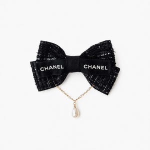 Peut inclure: Un accessoire de coiffure en forme de nœud en tweed noir, doté d'un ruban central noir portant l'inscription blanche CHANEL des deux côtés. Une fine chaîne dorée pend du nœud, retenant une perle fantaisie blanche en forme de goutte.
