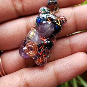 May include: A purple resin bear charm with a copper wire coil, amethyst crystal, and colorful glitter. The charm is attached to a black cord.