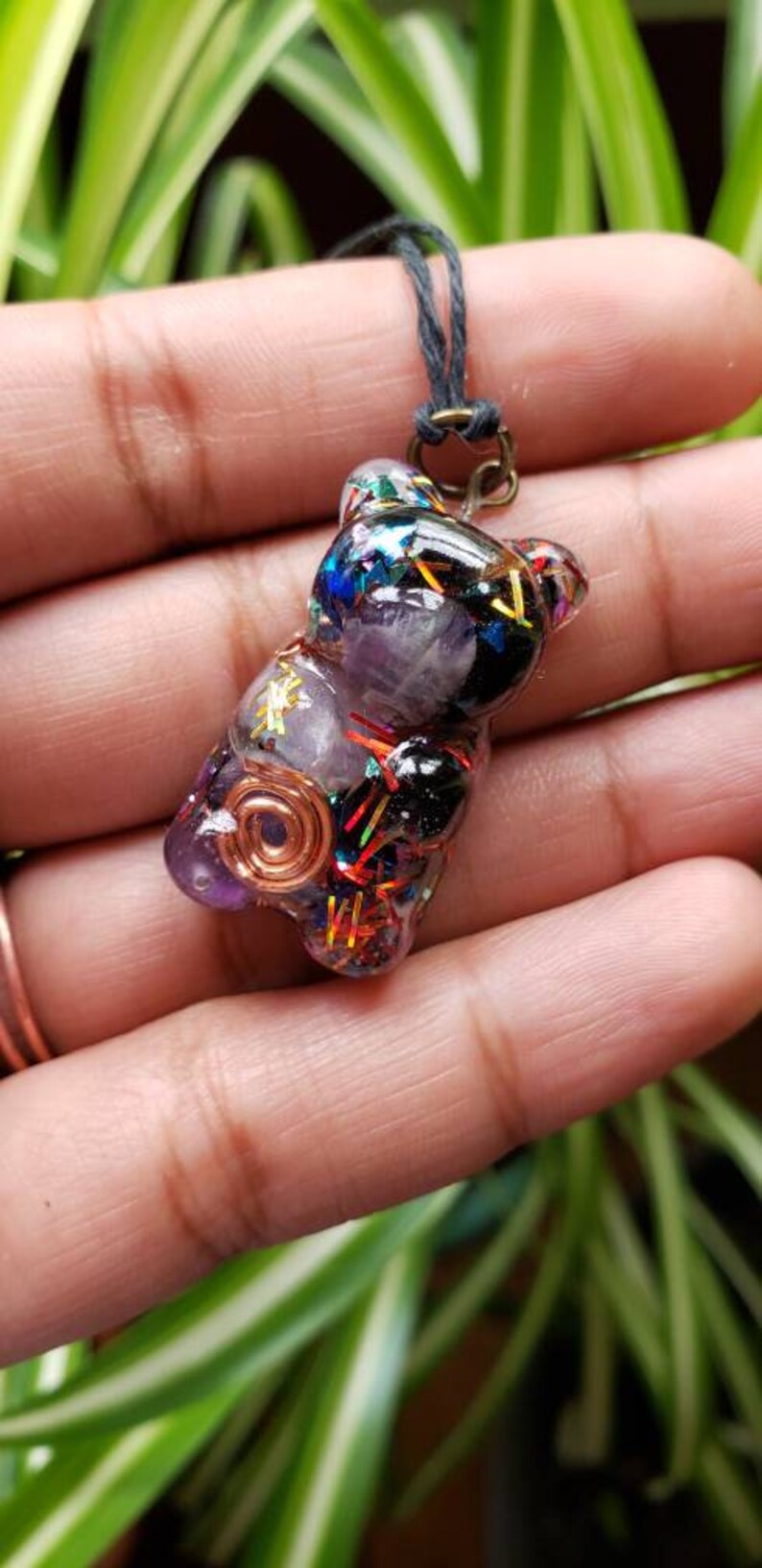 May include: A purple resin bear charm with a copper wire coil, amethyst crystal, and colorful glitter. The charm is attached to a black cord.