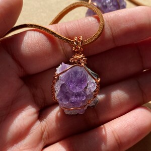 May include: A purple amethyst crystal wrapped in copper wire, hanging from a brown leather cord. The crystal is a cluster of smaller crystals, and the wire is twisted around the crystal to create a secure and decorative pendant.