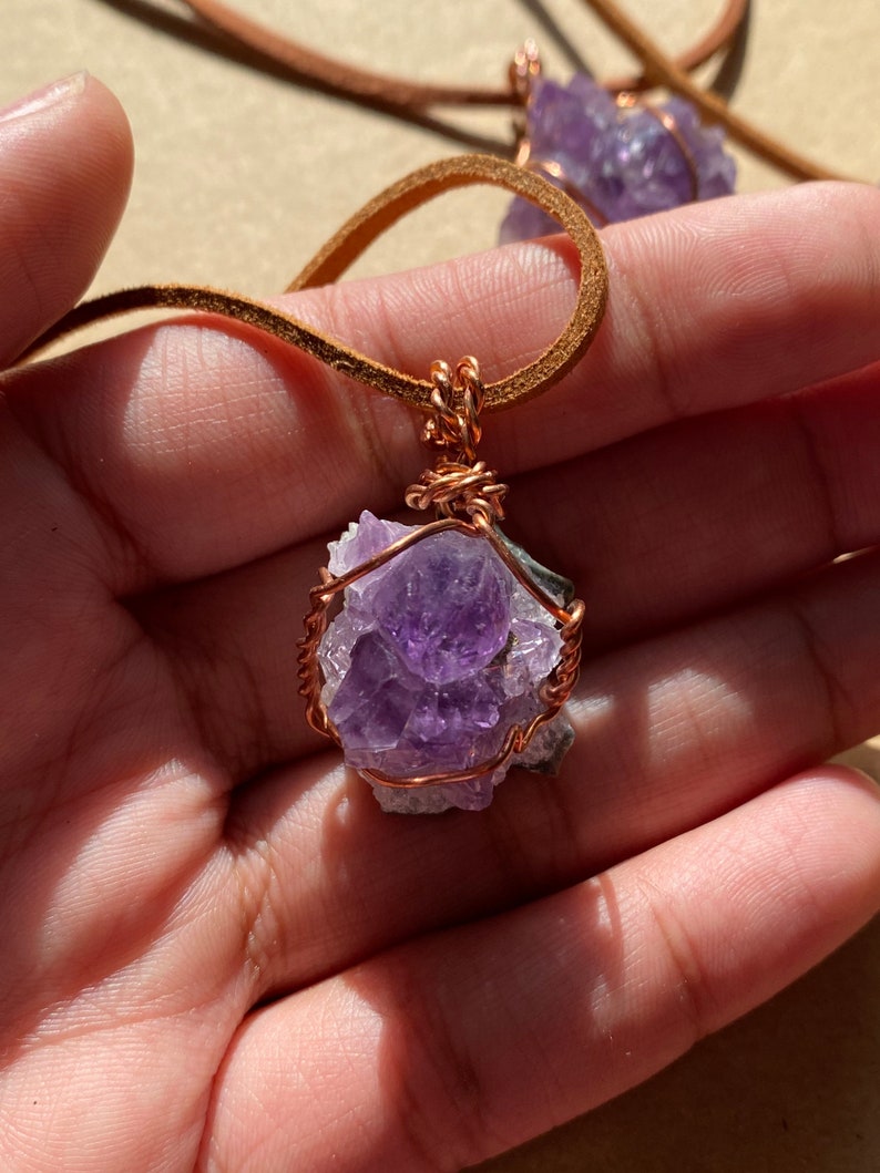 May include: A purple amethyst crystal wrapped in copper wire, hanging from a brown leather cord. The crystal is a cluster of smaller crystals, and the wire is twisted around the crystal to create a secure and decorative pendant.