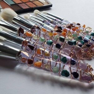 May include: A set of makeup brushes with handles wrapped in copper wire and various colourful gemstones. The brushes have white bristles and silver metal ferrules.