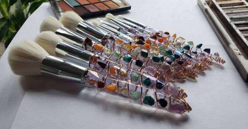 May include: A set of makeup brushes with handles wrapped in copper wire and various colourful gemstones. The brushes have white bristles and silver metal ferrules.