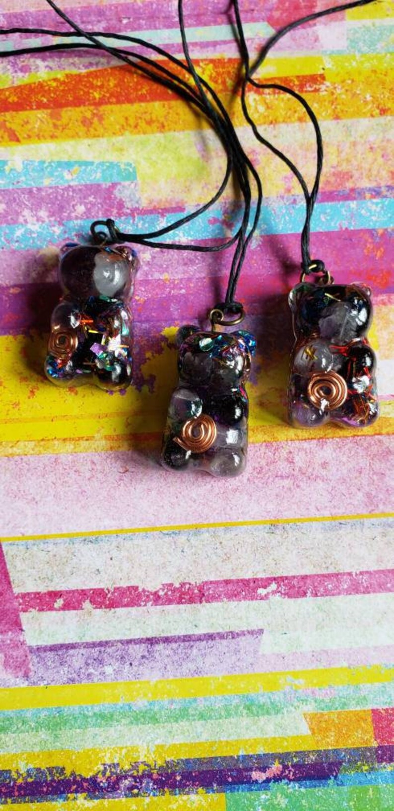 May include: Three resin bear pendants with a black and purple color scheme. Each pendant is filled with various crystals and a copper wire spiral. The pendants are hanging from black cords.