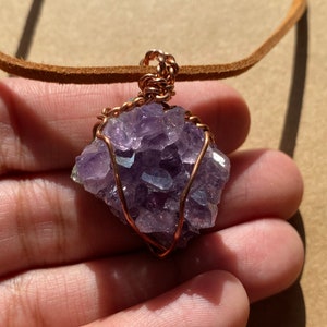 May include: A purple amethyst crystal wrapped in copper wire, hanging from a brown leather cord. The crystal is a cluster of small, faceted stones.