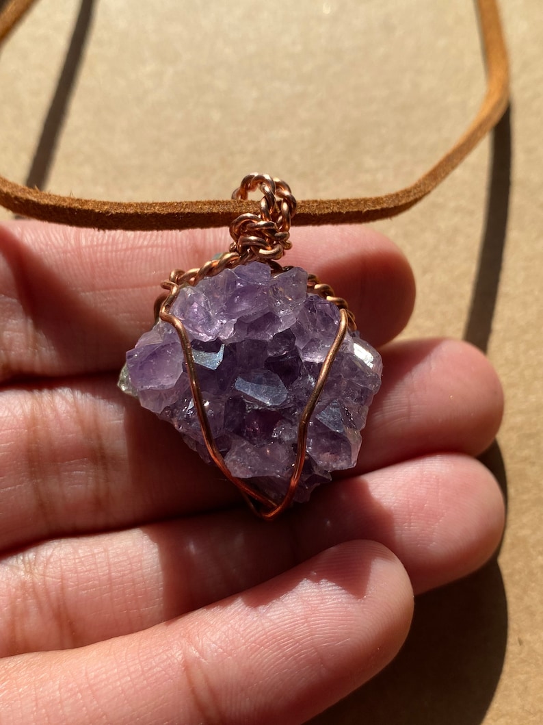 May include: A purple amethyst crystal wrapped in copper wire, hanging from a brown leather cord. The crystal is a cluster of small, faceted stones.
