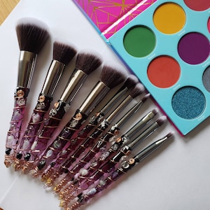 May include: Set of nine makeup brushes with purple and copper handles. The handles are decorated with clear and purple gemstones. The brushes have different sizes and shapes for various makeup applications.