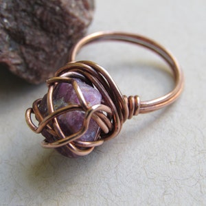 May include: A copper wire wrapped ring with a pink and white gemstone. The ring is on a white surface.