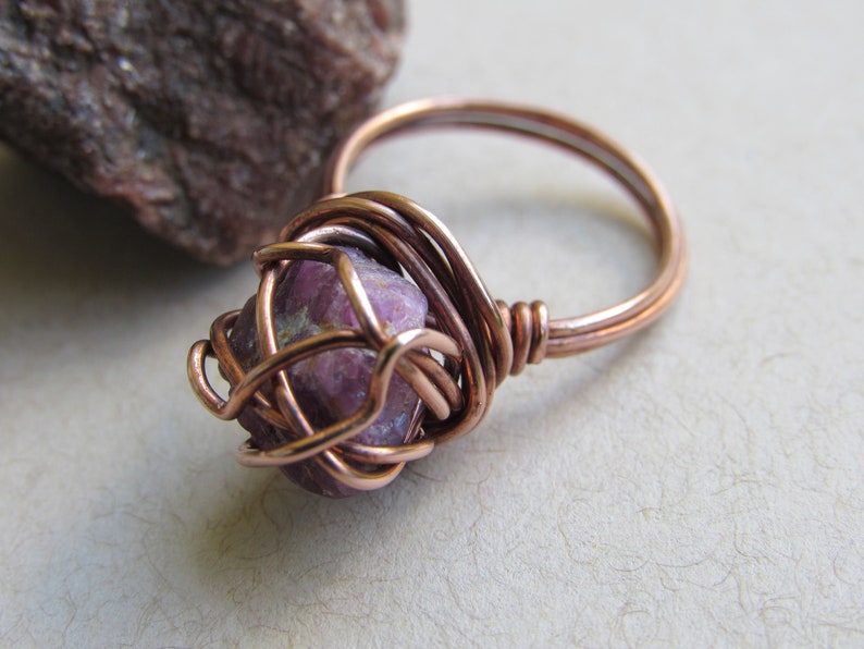 May include: A copper wire wrapped ring with a pink and white gemstone. The ring is on a white surface.