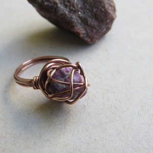 May include: A copper wire wrapped ring with a pink and white gemstone. The ring is on a white surface.