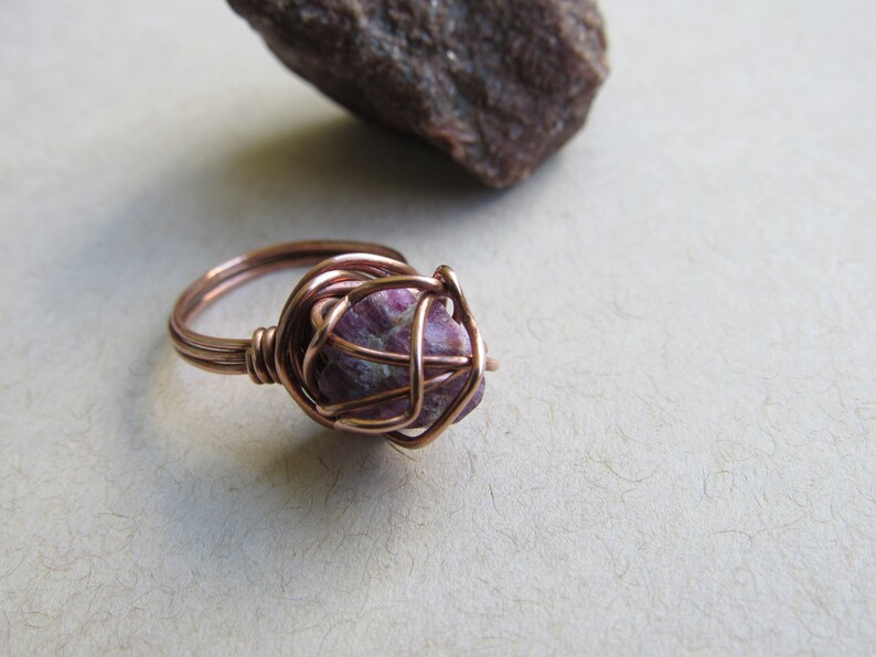 May include: A copper wire wrapped ring with a pink and white gemstone. The ring is on a white surface.
