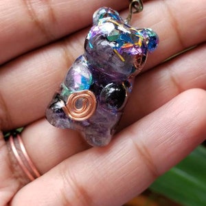 May include: A purple and black resin bear charm with iridescent glitter and copper wire accents. The charm is attached to a black cord.