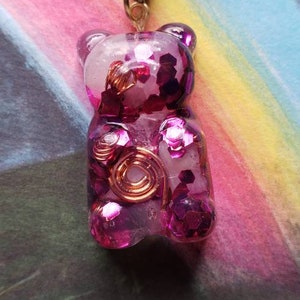 Op de afbeelding: Een ketting met een roze gummybeer-hanger met glitter en koperen draadaccenten. De hanger hangt aan een zwart koord.
