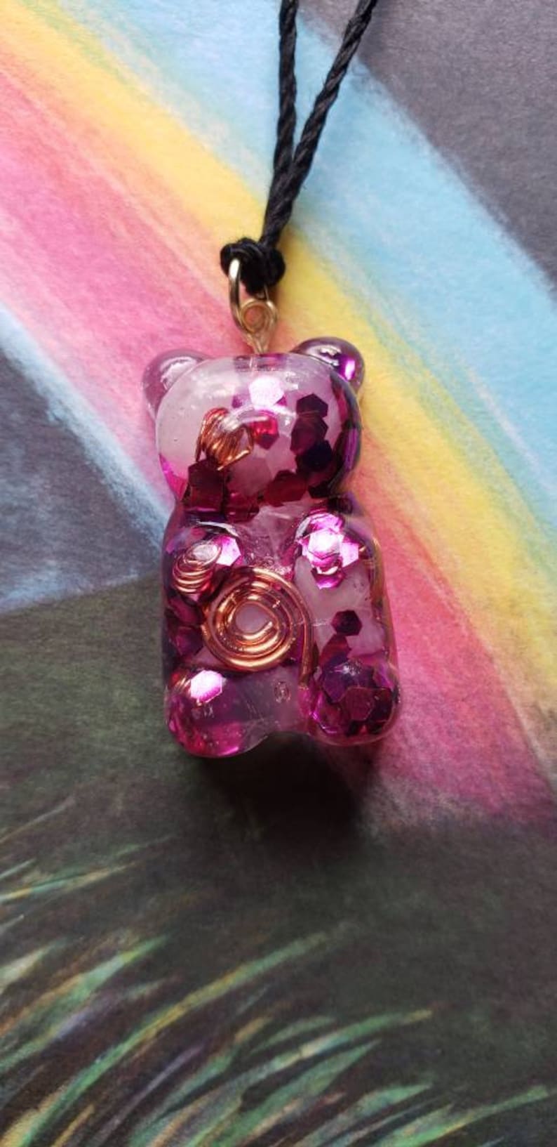 Op de afbeelding: Een ketting met een roze gummybeer-hanger met glitter en koperen draadaccenten. De hanger hangt aan een zwart koord.