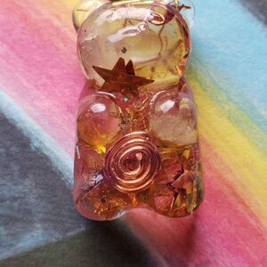 Op de afbeelding: Een gouden gummy bear hanger met helder en roze hars, met koperdraad en stervormige accenten. De hanger hangt aan een zwart koord.