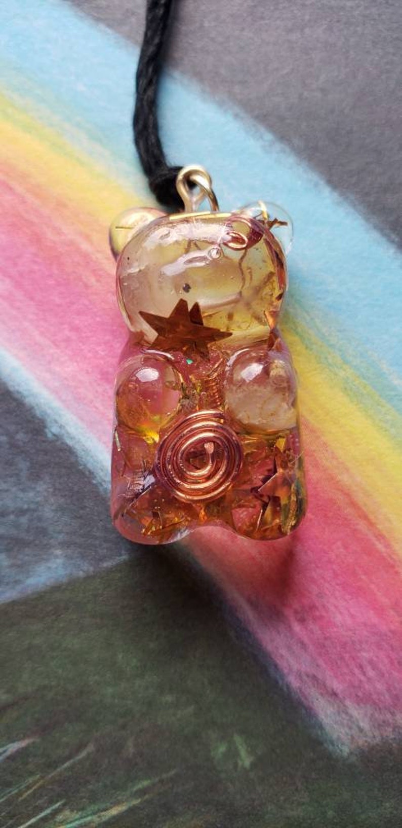 Op de afbeelding: Een gouden gummy bear hanger met helder en roze hars, met koperdraad en stervormige accenten. De hanger hangt aan een zwart koord.