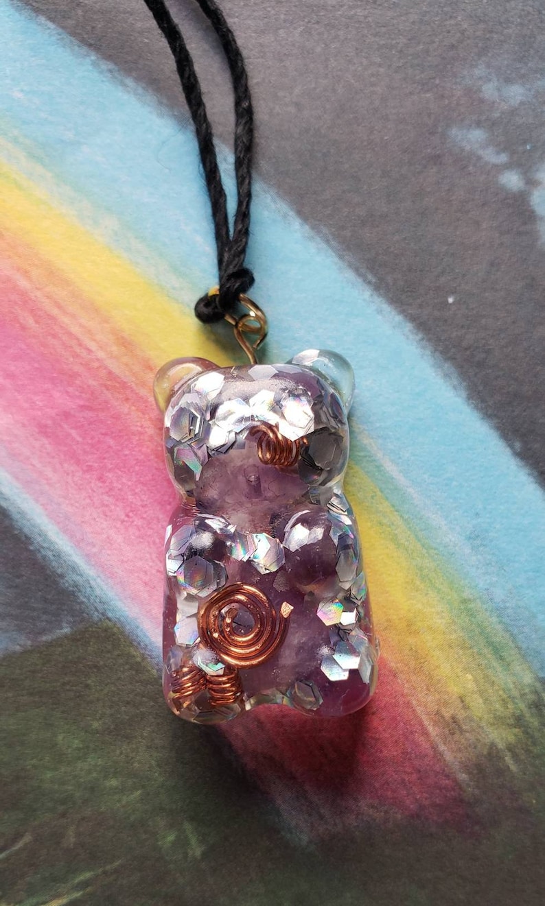 Op de afbeelding: Een paarse en zilveren glitterhars gummy bear hanger met koperen draadaccenten, hangend aan een zwart koord.