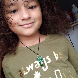 Op de afbeelding: Een jong persoon met krullend bruin haar draagt een groen shirt met gouden letters die "always be kind" zeggen en een groene gummy bear ketting.