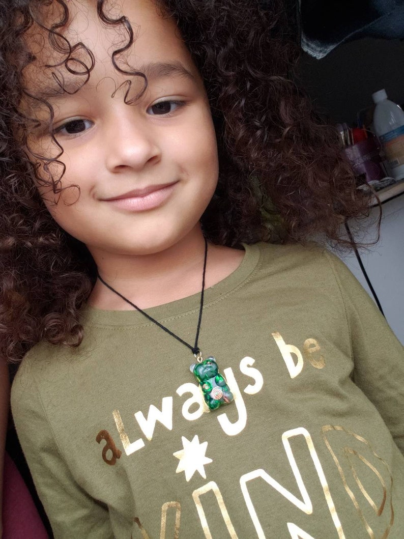 Op de afbeelding: Een jong persoon met krullend bruin haar draagt een groen shirt met gouden letters die "always be kind" zeggen en een groene gummy bear ketting.