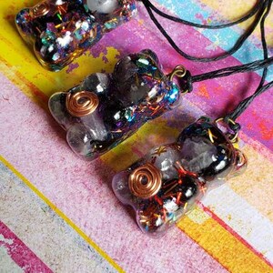 May include: Three resin bear pendants with black and clear crystal accents, copper wire, and iridescent glitter. The pendants are on a black cord and are displayed on a colorful striped background.